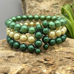 May include: Three beaded bracelets in shades of green and cream are stacked on a textured, brown surface. The bracelets are made of round beads, with a darker green accent bead design. Green leaves are visible in the background.