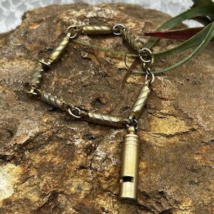 May include: A vintage gold-toned bracelet with a whistle charm. The bracelet features a series of textured, rectangular links and a small whistle pendant. The bracelet is displayed on a textured, brown surface.