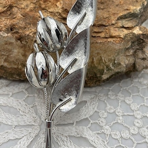 May include: A silver-toned brooch featuring a floral design with three tulip-shaped blooms and several textured leaves. The brooch has a stem-like base and is set against a textured background.