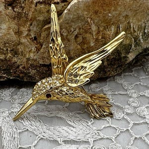 May include: A gold-toned hummingbird brooch featuring a textured metal finish on the wings and tail, with clear rhinestones embedded along the head and upper wing, and a single small dark red stone serving as the eye of the bird.