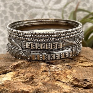 May include: A stack of silver-toned bangles with intricate detailing. The bracelets feature rows of small beads and decorative patterns. The jewelry is displayed on a textured, brown surface, with a blurred background.