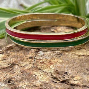 May include: Two stacked bangle bracelets made of gold-toned metal. The top bangle features a smooth, vibrant red enamel inlay, while the bottom bangle features a smooth, deep green enamel inlay, both set within the polished gold-toned metal frame.