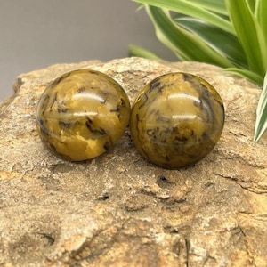 May include: Two round, decorative spheres with a marbled pattern in shades of yellow and brown. The spheres are resting on a textured, tan-colored surface, with green and white plant leaves in the background.