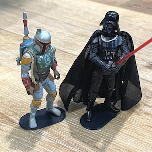 May include: Two Star Wars action figures, Darth Vader and Boba Fett, stand on black bases. Darth Vader is wearing a black cape and holding a red lightsaber. Boba Fett is wearing a green and brown suit and has a jetpack.