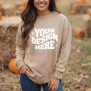 Könnte beinhalten: Beigefarbenes Sweatshirt mit dem weißen Text "YOUR DESIGN HERE". Das langärmlige Oberteil hat einen Rundhalsausschnitt und wird mit einer blauen Jeans getragen. Der Hintergrund zeigt Kürbisse und Heuballen.