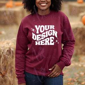 May include: A maroon crew neck sweatshirt with the white text "YOUR DESIGN HERE" and star details. The long-sleeved top is shown against a backdrop of hay bales and pumpkins, typical of autumn.