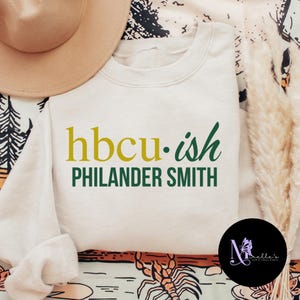 May include: A white sweatshirt with the text "hbcu.ish PHILANDER SMITH" in green and gold lettering.