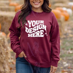 Może przedstawiać: Bordowa bluza z napisem "YOUR DESIGN HERE" w kolorze białym, z gwiazdkami. Bluza ma długie rękawy i okrągły dekolt. Modelka ma na sobie niebieskie dżinsy. Tło jest rozmyte z dyniami i sianem.