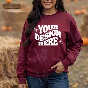 Könnte beinhalten: Ein weinrotes Sweatshirt mit dem Text "YOUR DESIGN HERE" in Weiß, mit Sternakzenten. Das Sweatshirt wird mit einer blauen Jeans getragen. Der Hintergrund umfasst Kürbisse und Heuballen, was auf ein Herbstthema hindeutet.
