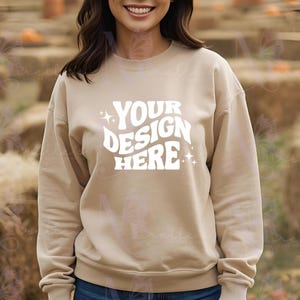 Może przedstawiać: Beżowa bluza z okrągłym dekoltem z białym napisem "YOUR DESIGN HERE" w stylu retro. Bluza z długim rękawem ma luźny krój. Tło jest rozmyte, co sugeruje otoczenie na zewnątrz.