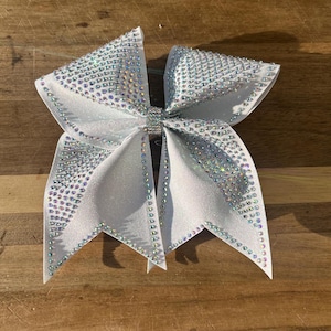 May include: A large white hair bow featuring a glittery fabric base and a dense covering of iridescent rhinestones. The bow is constructed with multiple loops and pointed tails, with a central band also encrusted with small clear crystals.