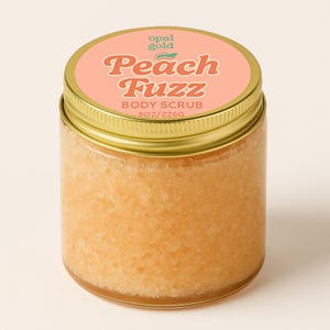 May include: A clear glass jar with a gold lid, filled with a peach-colored body scrub. The label reads "Peach Fuzz Body Scrub" in a retro font. The jar contains 8 oz of product.