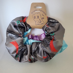 May include: A set of three grey, blue and purple satin scrunchies with a black and red design. The scrunchies have a tag attached that says "@manic_menagerie".