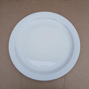 May include: A round, white ceramic plate with a slightly raised rim. The plate has a smooth, glossy surface and is set against a brown background. This simple, elegant plate is suitable for serving various dishes.