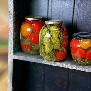 Può includere: Piccoli barattoli di verdure sottaceto, tra cui pomodori e cetrioli, sono esposti su una piccola mensola di legno. I barattoli hanno coperchi dorati e sono pieni di verdure ed erbe colorate. La mensola è dipinta in tonalità di grigio e verde.