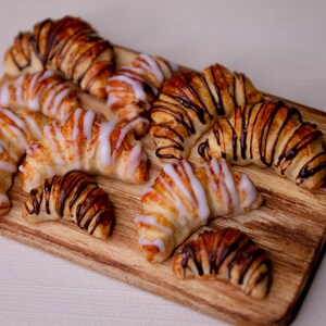 May include: A wooden board with a variety of croissants. Some are topped with white icing and others with chocolate drizzle.