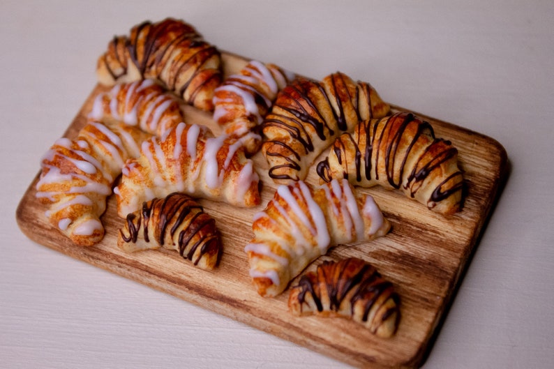 May include: A wooden board with a variety of croissants. Some are topped with white icing and others with chocolate drizzle.
