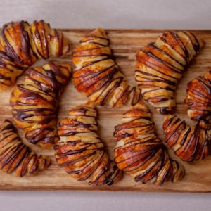 May include: A wooden cutting board with nine chocolate-covered croissants. The croissants are arranged in a semi-circle shape, with the tips of the croissants pointing towards the center of the board.