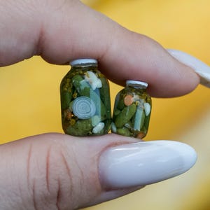 May include: Two miniature glass jars filled with pickles, onions, and carrots. The jars have white lids and are held between fingers against a yellow background. The pickles are green, and the onions are white.
