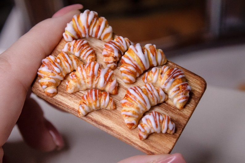 May include: A wooden board with nine miniature croissants, each with a white icing swirl. The croissants are arranged in a semi-circle on the board.