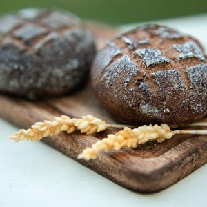 Op de afbeelding: Twee donkerbruine broden op een houten snijplank met twee tarwehalmen.