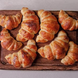 May include: A close-up of eight golden brown croissants arranged on a wooden cutting board. The croissants are slightly different sizes and shapes, but all have a flaky, buttery texture.