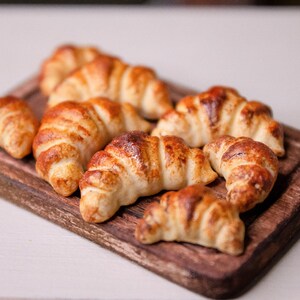 May include: A close-up of a wooden cutting board with eight golden brown croissants arranged on top. The croissants are slightly overlapping and have a flaky texture.