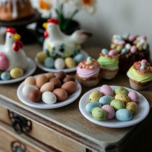 May include: A table setting with a variety of pastel-colored Easter eggs, cupcakes, and ceramic chickens. The eggs are arranged in three white dishes, while the cupcakes are decorated with pastel frosting and sprinkles. The ceramic chickens are white with pink accents.