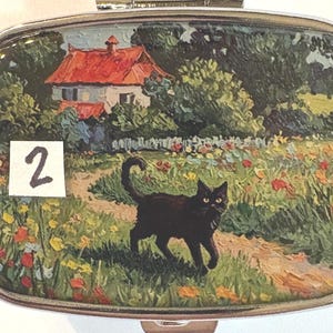 May include: A rectangular, silver-toned pill box with a painted landscape scene. The scene depicts a black cat walking on a path in a field of flowers, with a house and red roof in the background. The number "2" is on a white square.