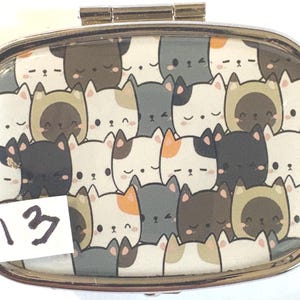 May include: A silver metal pill box with a hinged lid. The top features a pattern of cartoon cat faces in various colors, including white, gray, brown, and orange. A white sticker with the number "13" is on the bottom left.
