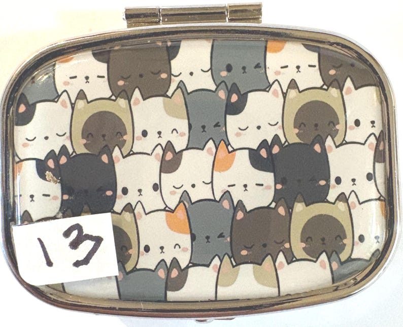 May include: A silver metal pill box with a hinged lid. The top features a pattern of cartoon cat faces in various colors, including white, gray, brown, and orange. A white sticker with the number "13" is on the bottom left.