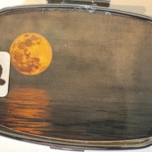 Puede incluir: Un pastillero plateado con tapa abisagrada. La caja presenta un gr&aacute;fico de luna llena y un reflejo sobre el agua. Una peque&ntilde;a pegatina blanca con el n&uacute;mero "12" est&aacute; adherida a la caja.