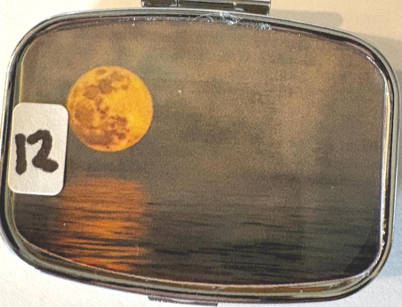 Puede incluir: Un pastillero plateado con tapa abisagrada. La caja presenta un gr&aacute;fico de luna llena y un reflejo sobre el agua. Una peque&ntilde;a pegatina blanca con el n&uacute;mero "12" est&aacute; adherida a la caja.