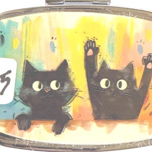 May include: A rectangular, silver-toned pill box with a hinged lid. The lid features a colorful illustration of two black cats with large eyes. One cat is waving. A white sticker with the number "15" is on the left.