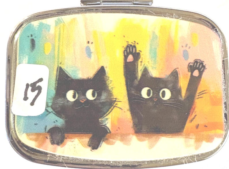 May include: A rectangular, silver-toned pill box with a hinged lid. The lid features a colorful illustration of two black cats with large eyes. One cat is waving. A white sticker with the number "15" is on the left.
