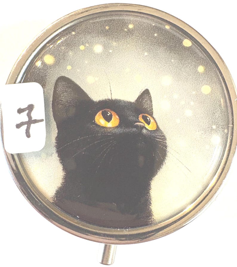 May include: A round, silver-toned pill box featuring a black cat with bright yellow eyes looking upwards. The background has a starry effect. A white label with the number 7 is on the left side of the box.