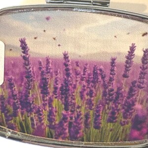 Puede incluir: Un pastillero plateado con un dise&ntilde;o de campo de lavanda. La imagen dentro de la caja presenta un vibrante campo de lavanda p&uacute;rpura bajo un cielo nublado, con peque&ntilde;os insectos voladores. Una peque&ntilde;a etiqueta blanca con el n&uacute;mero "II" est&aacute; a la izquierda.