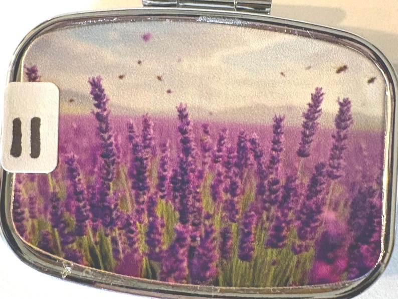Puede incluir: Un pastillero plateado con un dise&ntilde;o de campo de lavanda. La imagen dentro de la caja presenta un vibrante campo de lavanda p&uacute;rpura bajo un cielo nublado, con peque&ntilde;os insectos voladores. Una peque&ntilde;a etiqueta blanca con el n&uacute;mero "II" est&aacute; a la izquierda.