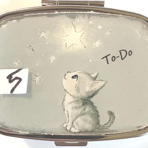 May include: A silver-toned pill box with a light blue background featuring a kitten looking up at a star. The text "To-Do" is written on the box. A white rectangle with the number "5" is in the lower left corner.