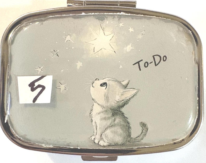 May include: A silver-toned pill box with a light blue background featuring a kitten looking up at a star. The text "To-Do" is written on the box. A white rectangle with the number "5" is in the lower left corner.