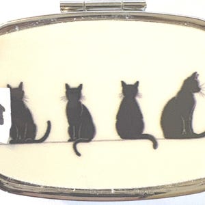May include: A silver pill box with a cream-colored background featuring four black cat silhouettes. The cats are sitting on a thin line. A white sticker with the number 9 is on the left side of the box.