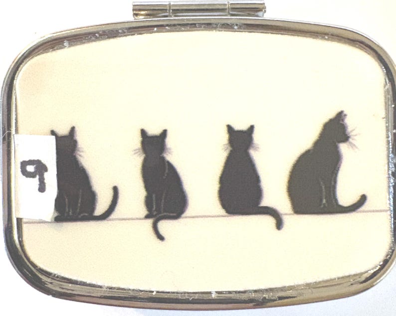 May include: A silver pill box with a cream-colored background featuring four black cat silhouettes. The cats are sitting on a thin line. A white sticker with the number 9 is on the left side of the box.