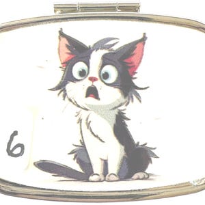 May include: A silver-toned pill box with a hinged lid. The box features a cartoon illustration of a surprised black and white cat with large blue eyes and pink ears. The number "6" is printed in black on the left side of the box.