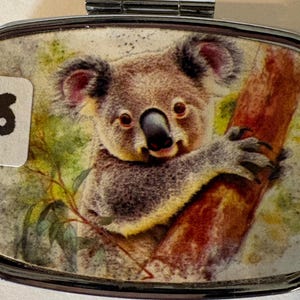 Puede incluir: Un pastillero rectangular plateado con tapa abisagrada. La tapa presenta una imagen estilo acuarela de un koala aferrado a una rama de &aacute;rbol. Una peque&ntilde;a pegatina blanca con el n&uacute;mero "3" est&aacute; adherida a la caja.