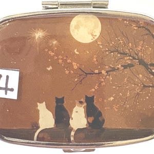 May include: A silver pill box with a decorative scene featuring four cats silhouetted against a full moon and flowering tree. The number "4" is in the lower left corner. The background is a warm brown color.