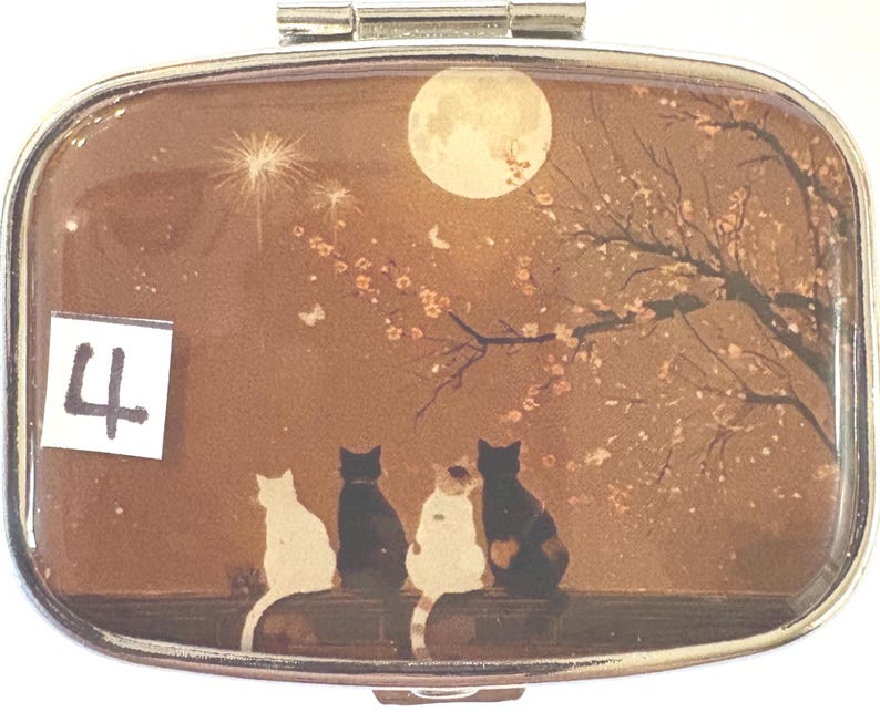 May include: A silver pill box with a decorative scene featuring four cats silhouetted against a full moon and flowering tree. The number "4" is in the lower left corner. The background is a warm brown color.