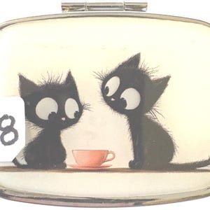 May include: A rectangular, silver-toned compact mirror with a whimsical illustration. Two black cartoon cats with large eyes are depicted, facing each other near a pink teacup. The number "8" is visible on the left. The background is off-white.