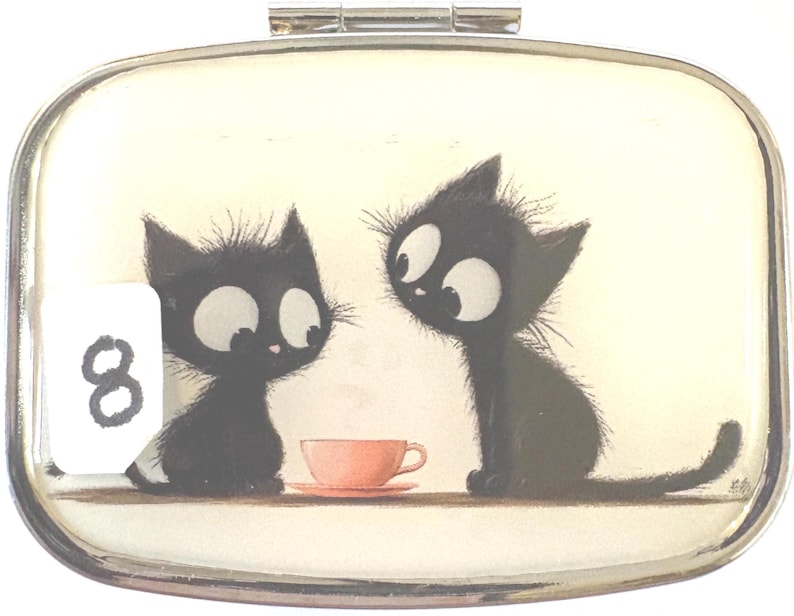May include: A rectangular, silver-toned compact mirror with a whimsical illustration. Two black cartoon cats with large eyes are depicted, facing each other near a pink teacup. The number "8" is visible on the left. The background is off-white.