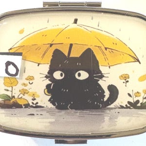 May include: A rectangular, silver-toned pill box with a hinged lid. The lid features a cartoon illustration of a black cat under a yellow umbrella in the rain. The number "10" is on a white sticker in the upper left corner.