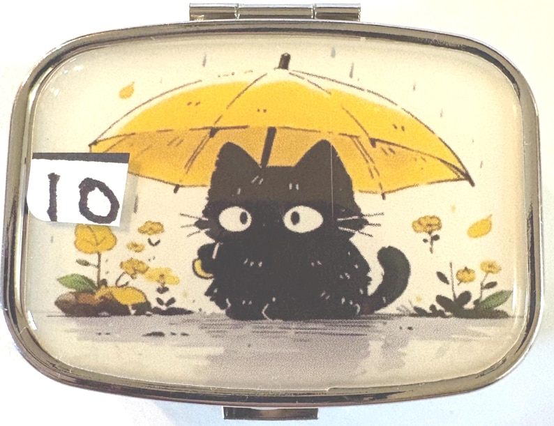 May include: A rectangular, silver-toned pill box with a hinged lid. The lid features a cartoon illustration of a black cat under a yellow umbrella in the rain. The number "10" is on a white sticker in the upper left corner.